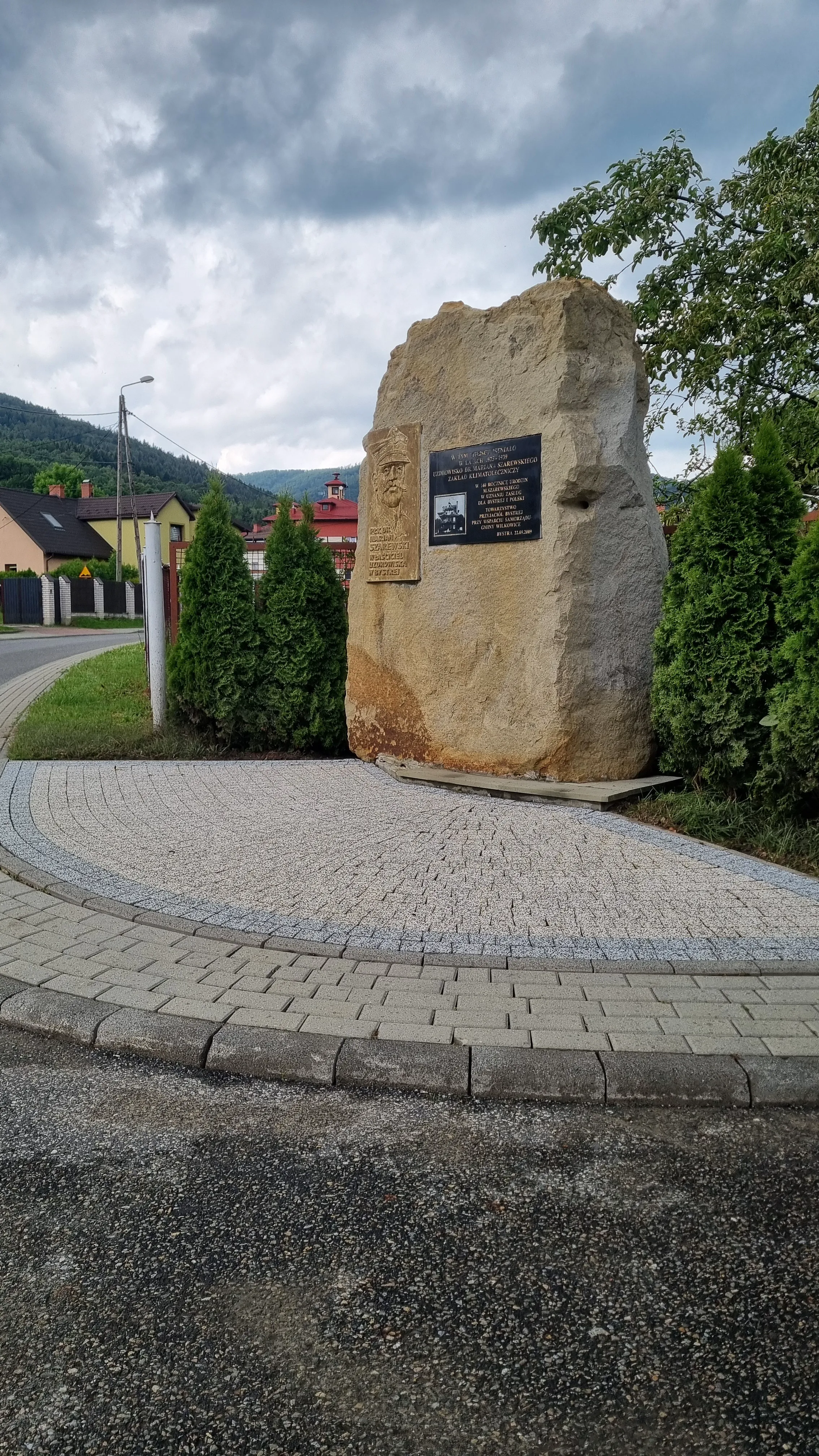 Pomnik w formie kamienia z tablicą na tle zieleni, przy drodze