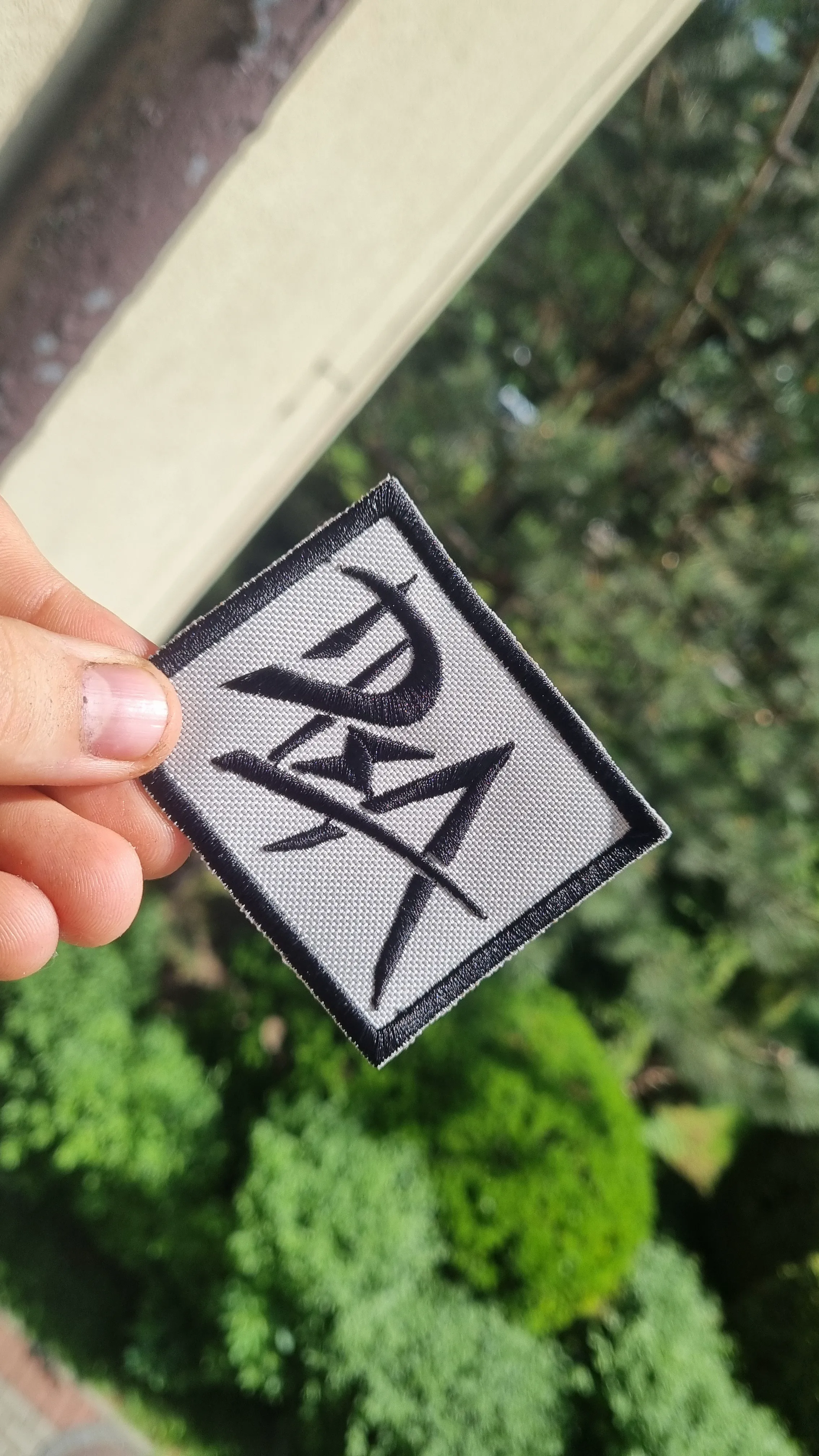 Naszywka z logotypem, trzymana ręką na tle zielonych drzew