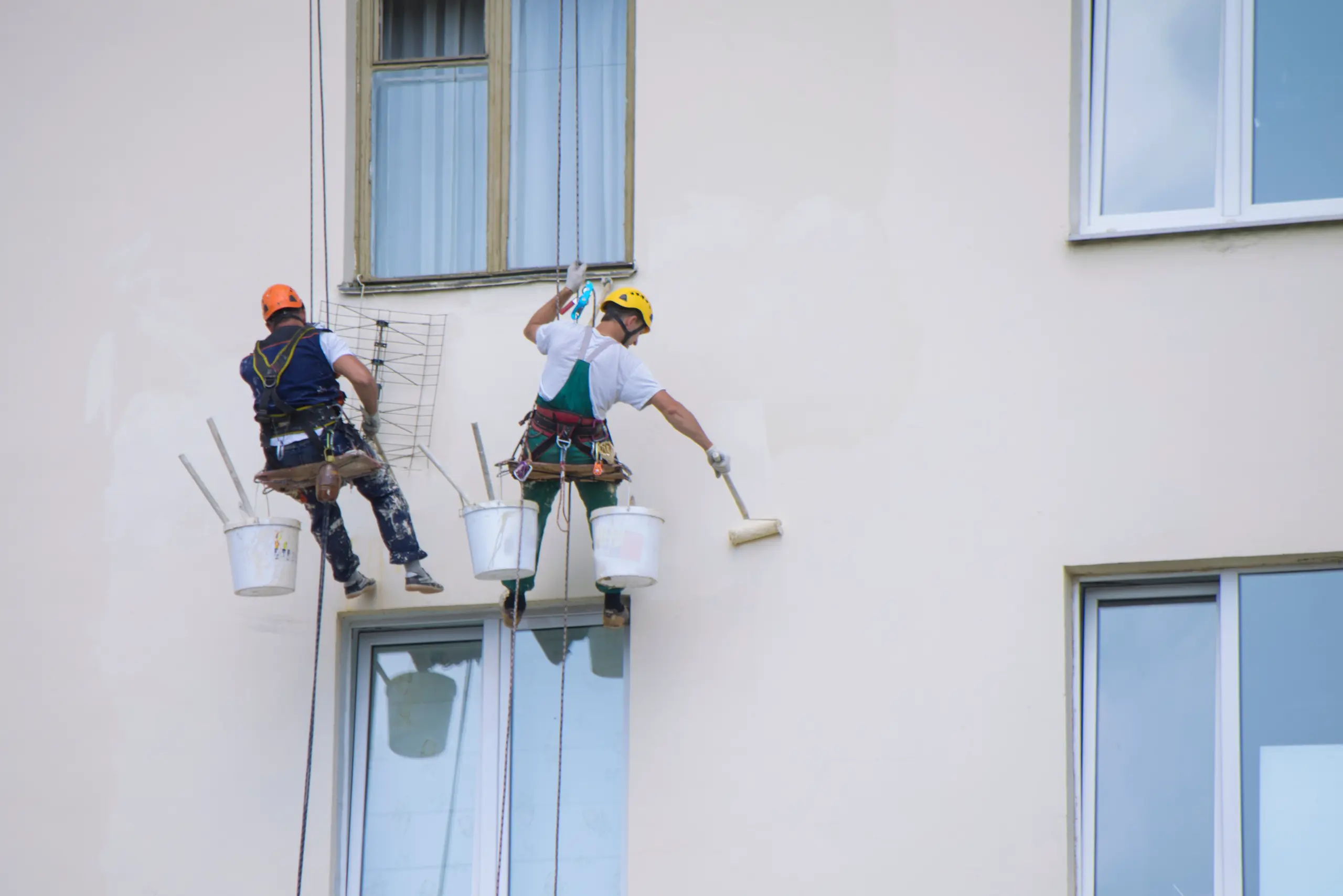 Pracownicy malują elewację budynku z użyciem wałków w technice alpinistycznej